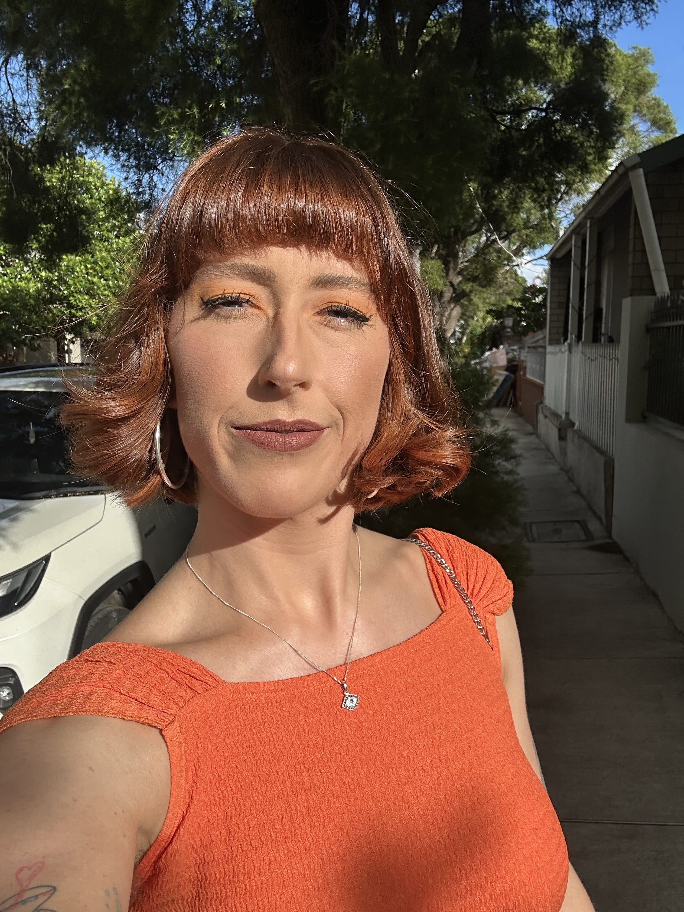 Portrait of a woman outdoors wearing an orange textured top and a pendant necklace, with trees and a sidewalk in the background.