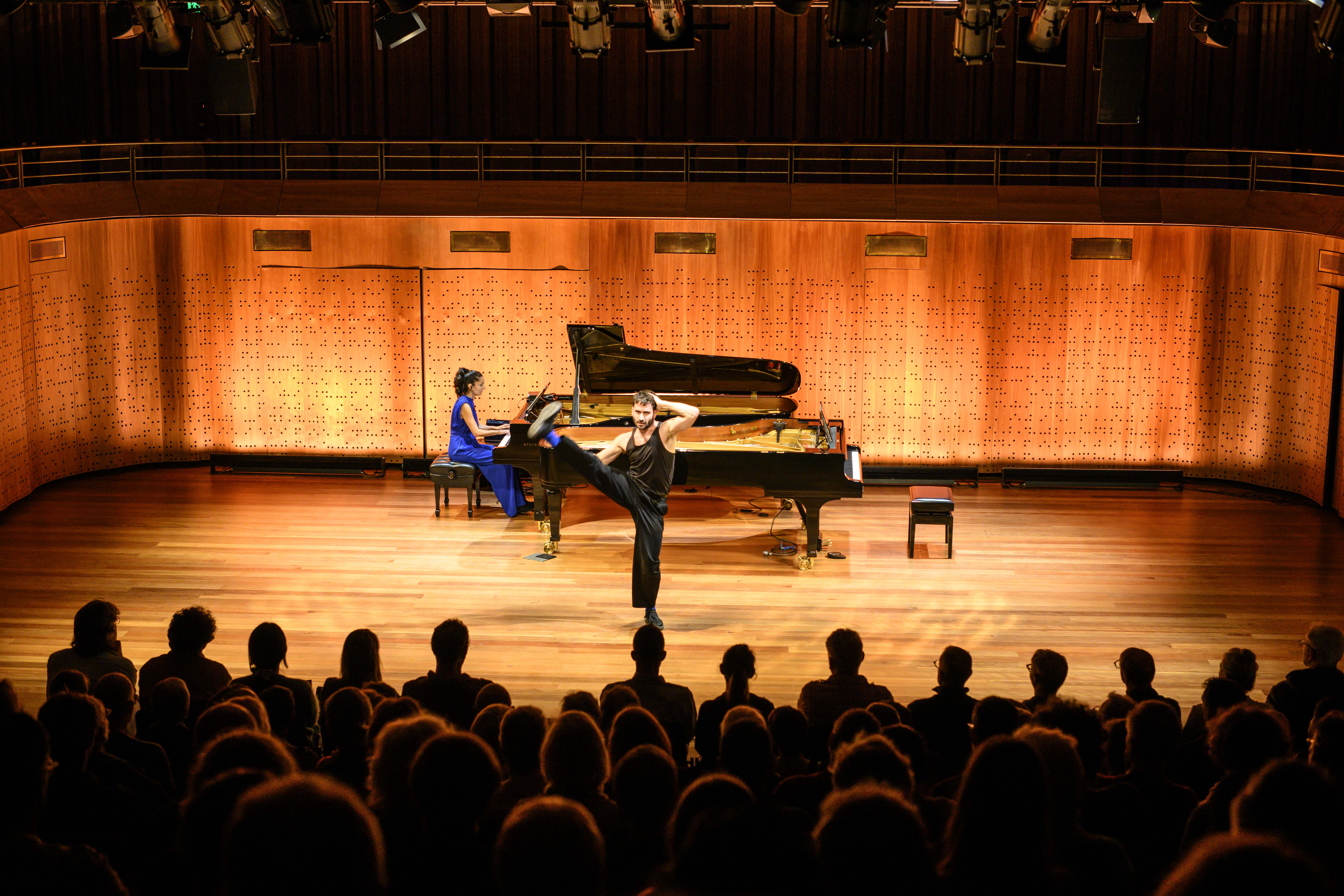 Pianist in a blue dress plays a grand piano onstage while a male dancer in black performs a high kick in front of an audience.