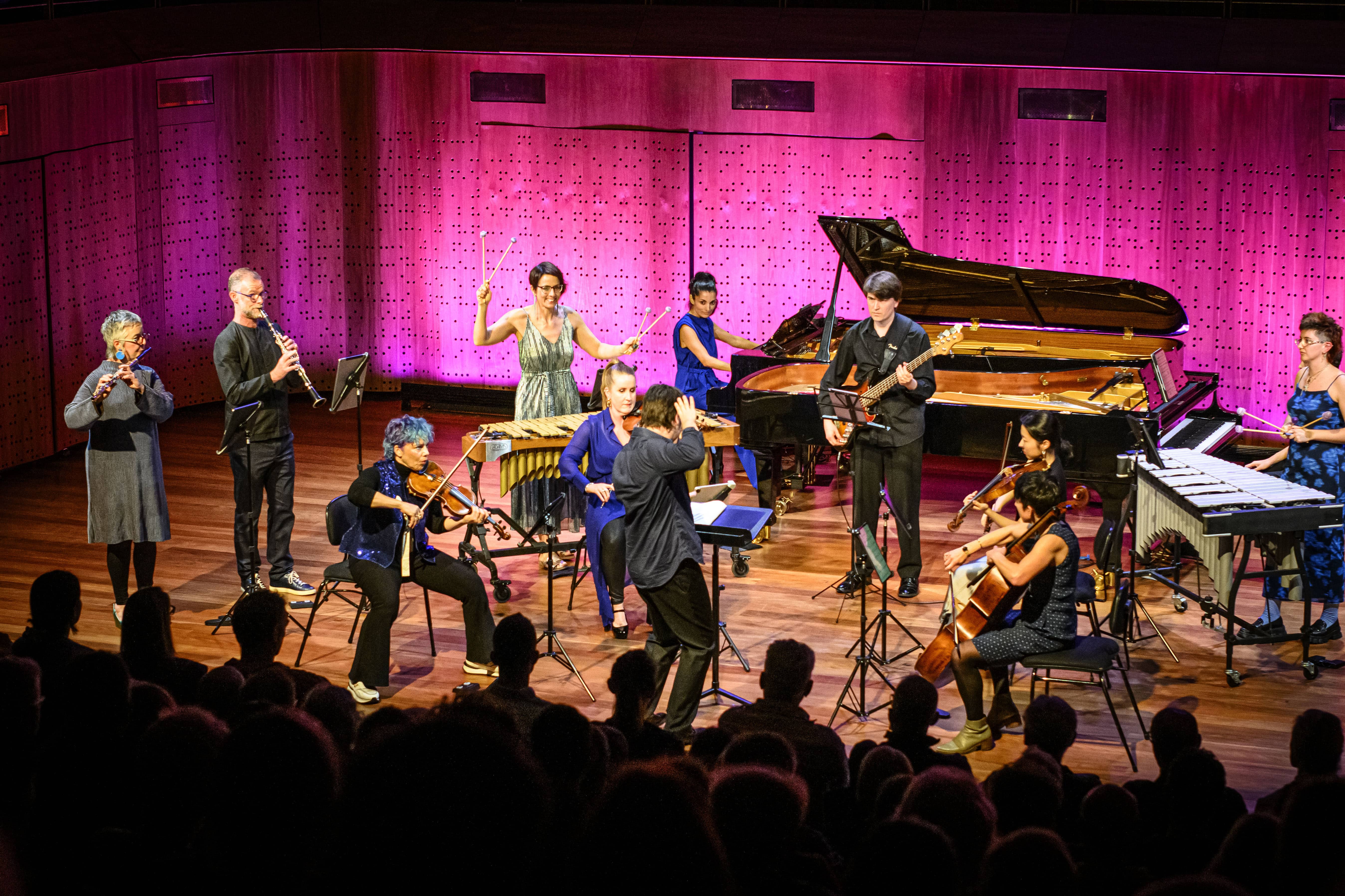 Orchestra on stage performing live; musicians play violin, piano, bass, percussion, and strings under magenta lighting with an audience in the foreground.