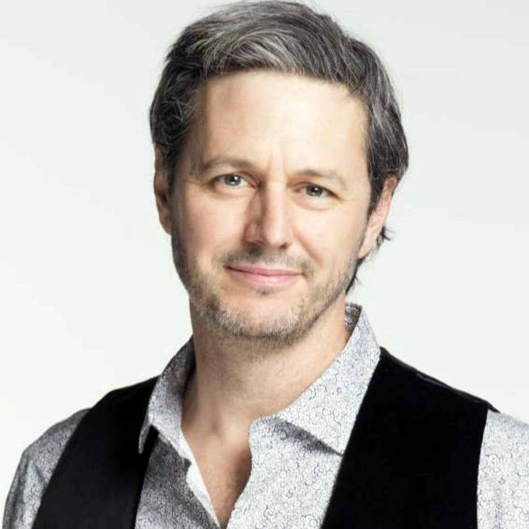 Smiling man with salt-and-pepper hair wearing a silver patterned shirt and black vest.
