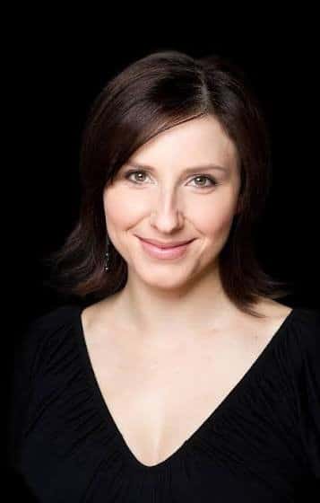 Portrait of a smiling woman with shoulder-length brown hair wearing a black V-neck top, against a black background.